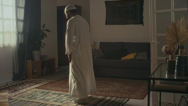 Rear wide footage of unidentified religious man in white cap and thawb making ruku and taking sujud position while expressing gratitude to Allah on prayer mat at home