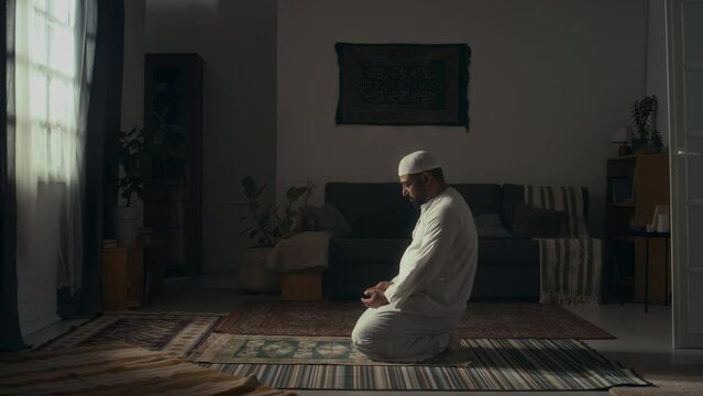 Side wide footage of bearded muslim man in white sitting alone on floor in spacious room and praying takbirs while reciting Koran at home