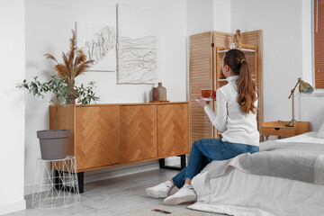 Young woman with cup of coffee looking at 3D paintings in bedroom, back view