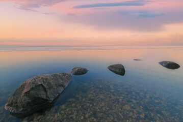 Tranquil sunset landscape at lake coast with a pink warm glow reflecting in water and snow and large rocks - 750934230
