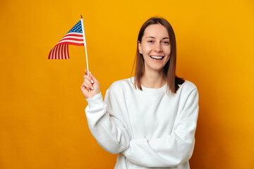 Ecstatic young smiling woman is holding a USA flag over yellow background. Studio portrait.