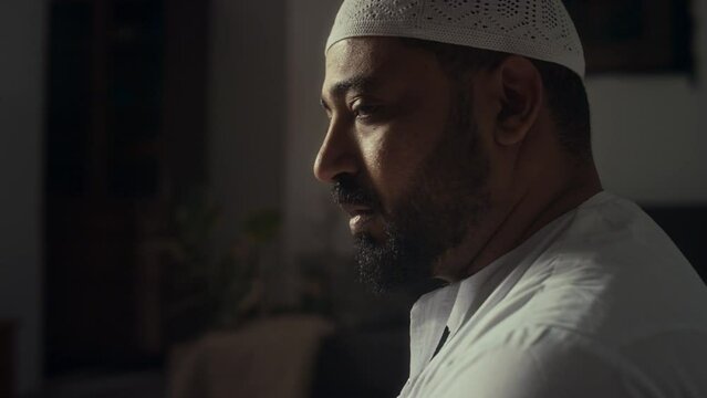 Side closeup of bearded Biracial muslim in white cap performing namaz at home while turning head to right and left