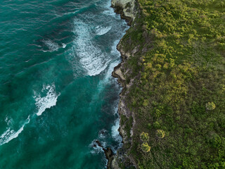 Aerial. Dark turquoise water with white foamy waves and green rocky shore
