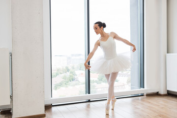 Young Caucasian ballerina in white tutu dancing on pointe in large bright modern hall in front of...