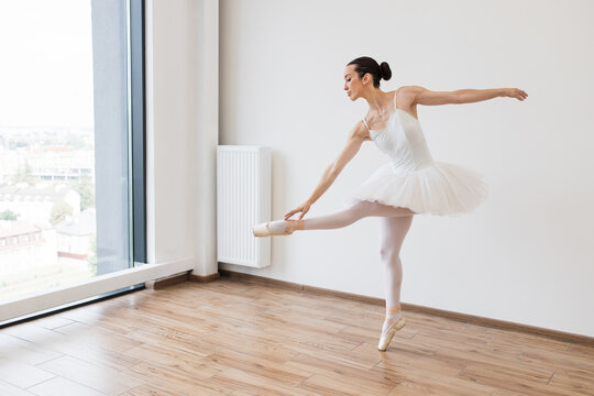 Young slender Caucasian ballerina in white tutu in pointe shoes dancing in large beautiful white hall. Graceful classical female dancer stretching her legs.