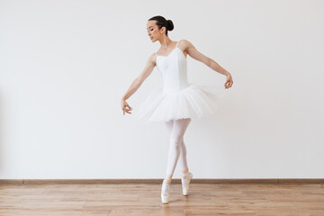 Beautiful young female dancer posing on studio background. Caucasian ballerina in white bodysuit and tutu poses in motion showing ballet elements while standing on pointe shoes.