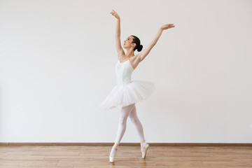 Caucasian young woman ballerina in white tutu, dancing on pointe with arms overhead, in the studio against a light bright background of modern studio hall.