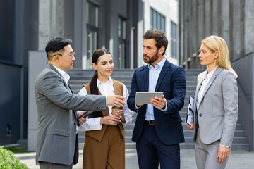 A group of business professionals engaged in a discussion outside office buildings. They are dressed smartly in suits, and one is holding a digital tablet.