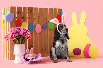 Cute cocker spaniel in bunny ears with Easter cake, paper eggs, rabbit, beautiful tulips and wooden fence on pink background