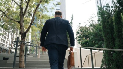 Back view of caucasian manager with formal suit walking up stairs while holding his suitcase in green city. Professional business man going to workiplace while climb stair in eco urban city. Urbane..