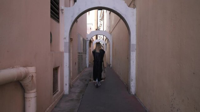 A woman in formal wear is strolling through a narrow alleyway with art on the building facades. Her sleeve grazes the wall as she gestures towards a door with a unique font
