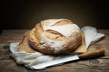 Whole loaf of traditional bread with knife, cutting board and tea towel on old wooden table, space for text. - 750965860