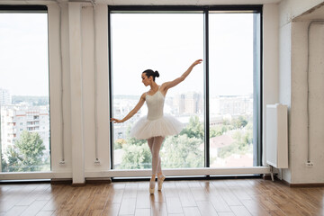 Beautiful graceful ballerina practice ballet positions in white tutu skirt near large window in...