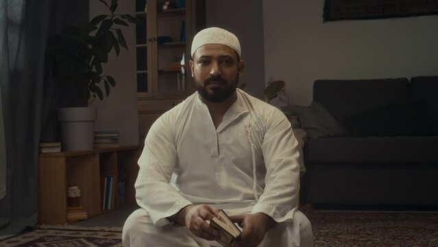 Medium handheld portrait of Biracial bearded muslim man in white skullcap and kandora sitting and posing with mini holy Quran book at home and looking at camera