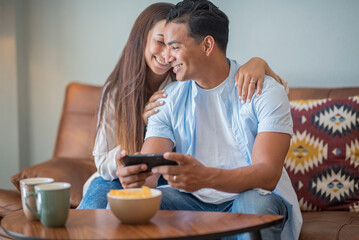Young couple watching online content in a smart phone sitting on a sofa at home in the living room....
