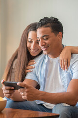 Portrait of cheerful positive young lovely couple smiling spending time together at home sitting on sofa typing on smartphone, searching internet using social network app on cellphone, family concept