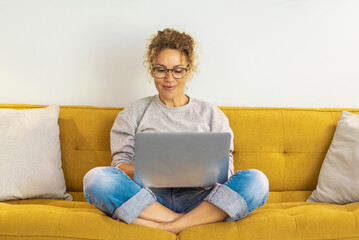 One woman smiling and using laptop computer at home sitting comfortably on a yellow sofa in living...