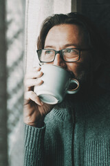 One man with thoughtful and serene expression on face drinking coffee or tea alone at home looking...