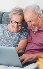 Aged couple at home paying bills on line with laptop and laughing a lot having fun together. Happiness and elderly lifestyle. Man and woman old senior sitting on sofa with computer online connection