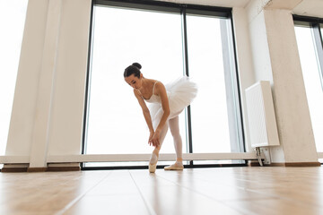Young graceful ballet dancer rehearsing performance in white hall room. Caucasian ballerina stretching to feet while standing in dance studio with large windows.