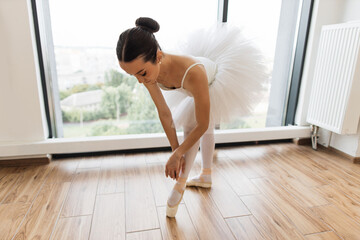 Beautiful graceful ballerina practice ballet positions in tutu skirt near large window in white...
