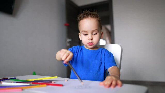 Charming baby boy sitting at desk holding a pencil. Cute kid drawing and talking. Blurred backdrop.