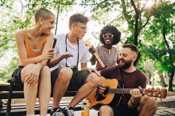 Group of multicultural friends singing and playing a guitar in park.