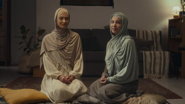Full handheld portrait of two pretty muslim ladies wearing hijabs and abayas posing while sitting on pillows in floor in living room and looking at camera