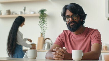 Happy bearded man homeowner husband sit in kitchen muslim indian guy smiling cheerful smile while woman wife cooking breakfast household work culinary. Married Arabian ethnic couple cook food at home