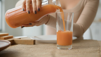 Close up unrecognizable woman pour orange juice from jar to glass on kitchen table. Female hands...