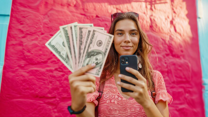 Candid shot of a young brunette holding a wad of banknotes while taking a selfie with her cell phone - 750988262