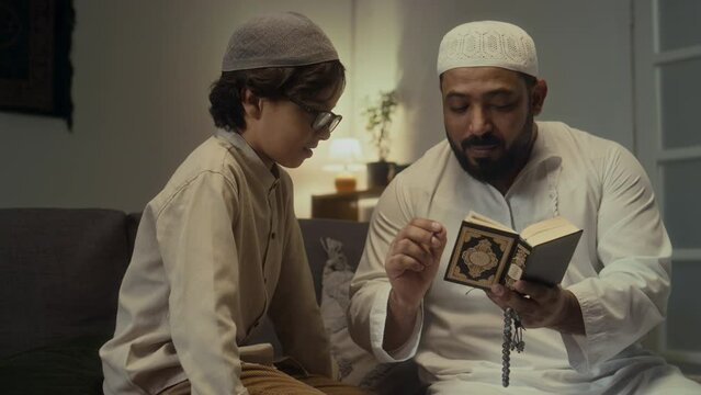 Medium footage of attentive little boy learning phrases for praying namaz while repeating after father who reciting Quran sitting in evening at home