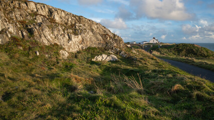 Irish landscape presents peninsula, grass, rocks, sea water, couple of white buildings and tourists