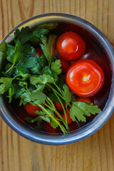 Fresh vegetables are being washed  in the metal bowl filled with cold water on the vintage cracked wooden cutting board. Organic tomatoes, parsley, celery. Top view. Copy space