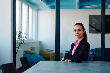 Successful young female leader in a suit with a pink shirt sitting in a modern glass office with a determined smile.