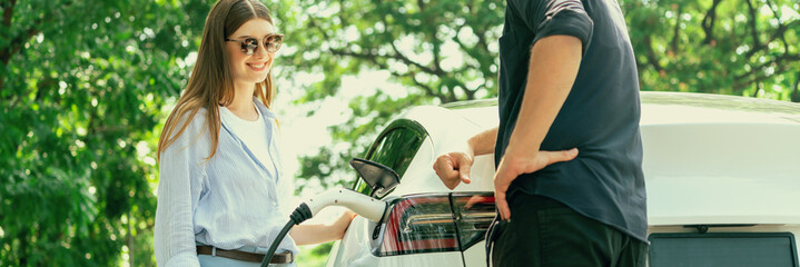 Focused EV car recharging electricity for battery on blurred background of lovey couple ruing their...