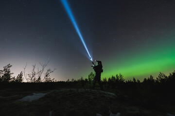 A soldier with a rifle and weapon flashlight in a night forest, sky with stars and northern lights.