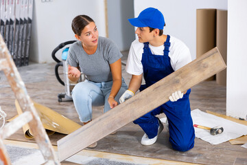 Interested young woman supervising laminate flooring installation in her apartment during renovation, talking to builder