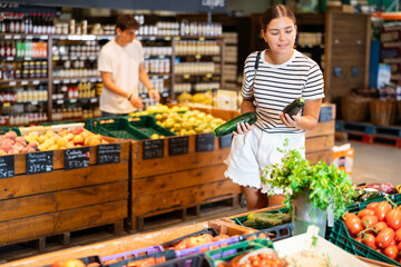 Positive young female shopper making purchases in supermarket, looking with interest for ripe...