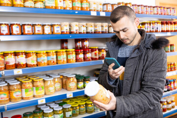 Adult male shopper with mobile phone choosing sauerkraut in grocery store