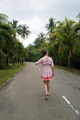 View from behind on a young caucasian woman wearing a pink dress walking on the road with the dog in a tropical setting
