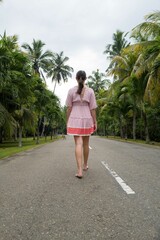 View from behind on a young caucasian woman wearing a pink dress walking on the road in a tropical setting