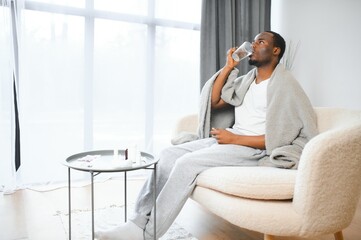 Serious young black bearded male sitting on bed in bedroom, hold glass of water and tablet on white...