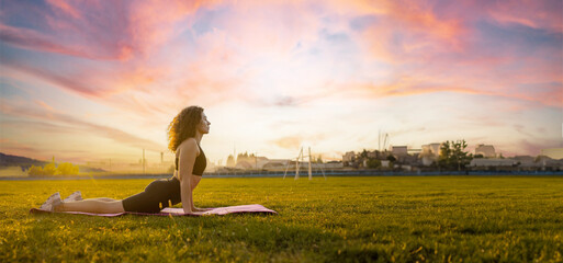 athletic curly woman in sportswear stands in a plank on a gymnastic mat in the morning at dawn on the grass in a field near the city.