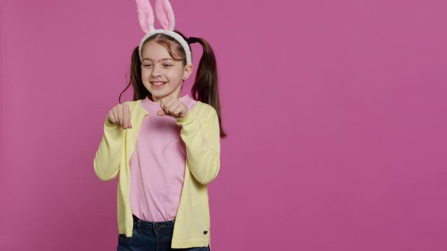Joyful carefree schoolgirl jumping around in studio, imitating a rabbit and hopping against pink background. Cheerful active child wearing bunny ears and bouncing, adorable kid. Camera A.