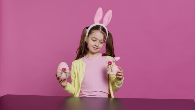 Small cheerful girl showing colorful decorated ornaments on camera, creating handcrafted festive arrangements. Young child feeling excited about easter sunday, holds rabbit and egg. Camera A.
