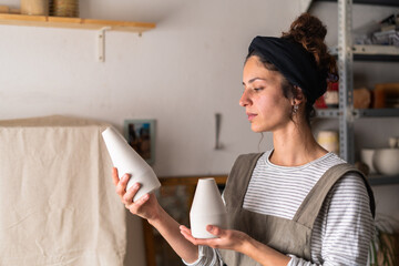 A focused female ceramist examines the shape and size of two unfinished ceramic vases in her...