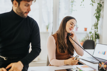 Professional woman multitasking at work with phone and laptop, colleague in background, in a bright...