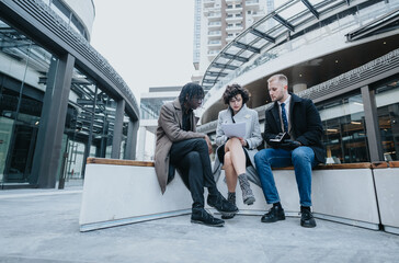 Three business employees collaborate on a project while seated outdoors in an urban setting, reviewing content on a laptop.