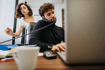Busy office scene with a man multitasking on a call and computer, and a woman in the background.